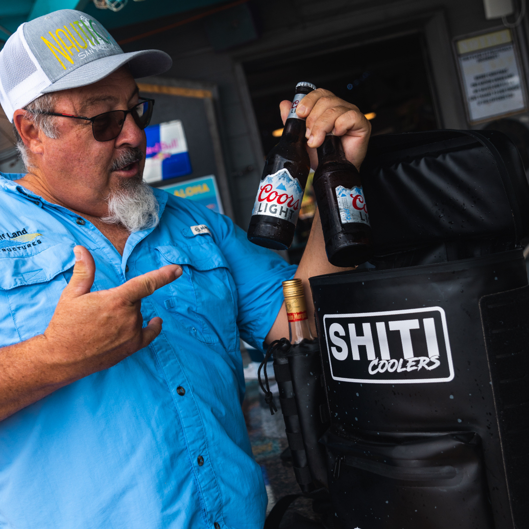 Man holding two beer bottles next to a Blackout Backpack Cooler, showcasing its capacity for 15 cans.