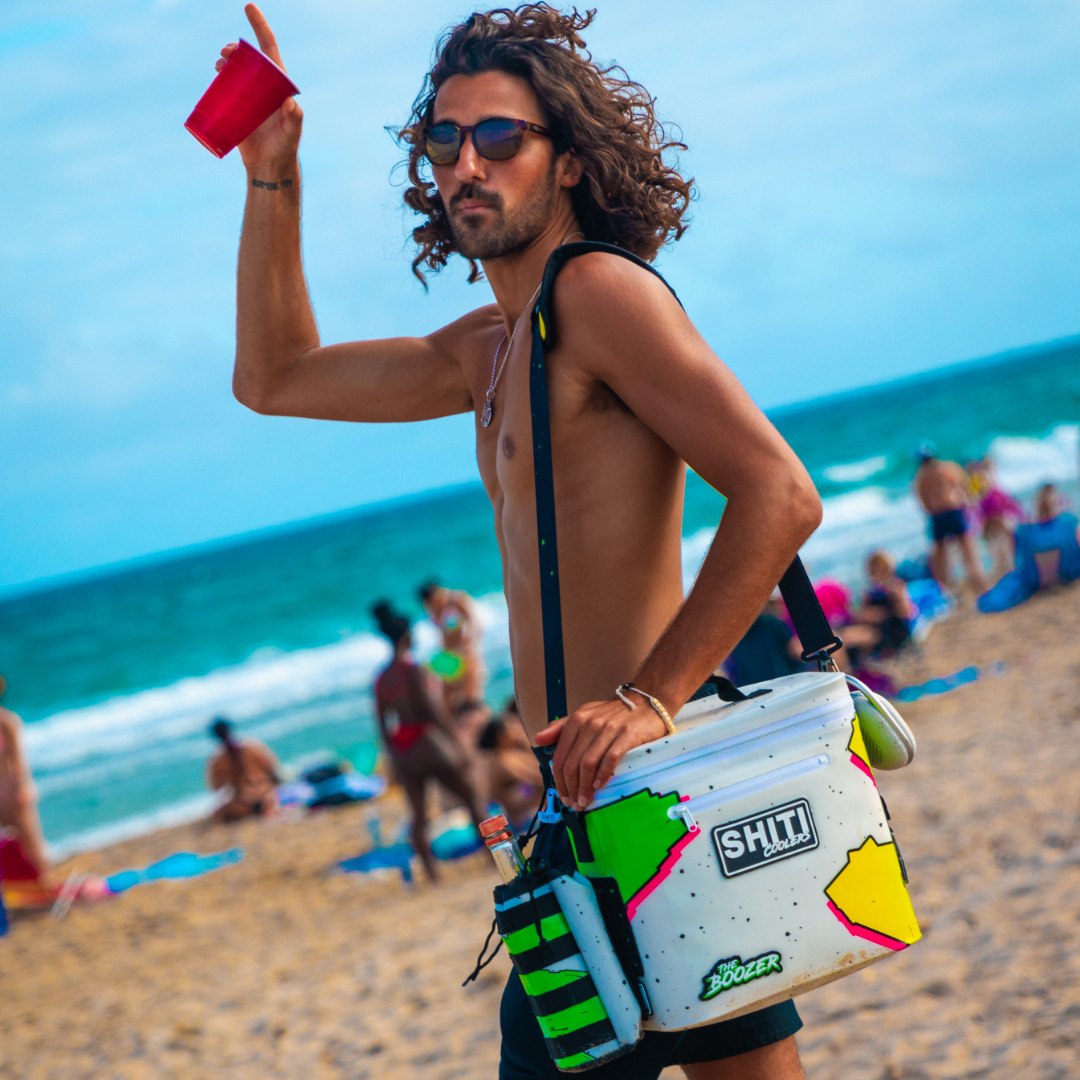 A man at the beach holds a red cup while carrying a colorful Boozer Soft Cooler, surrounded by beachgoers and ocean waves.