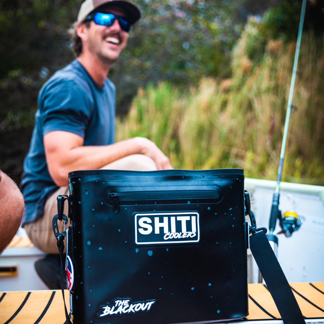 Blackout Soft Cooler by SHITI Coolers, designed to hold 12 cans, sits on a boat with a smiling man in the background.