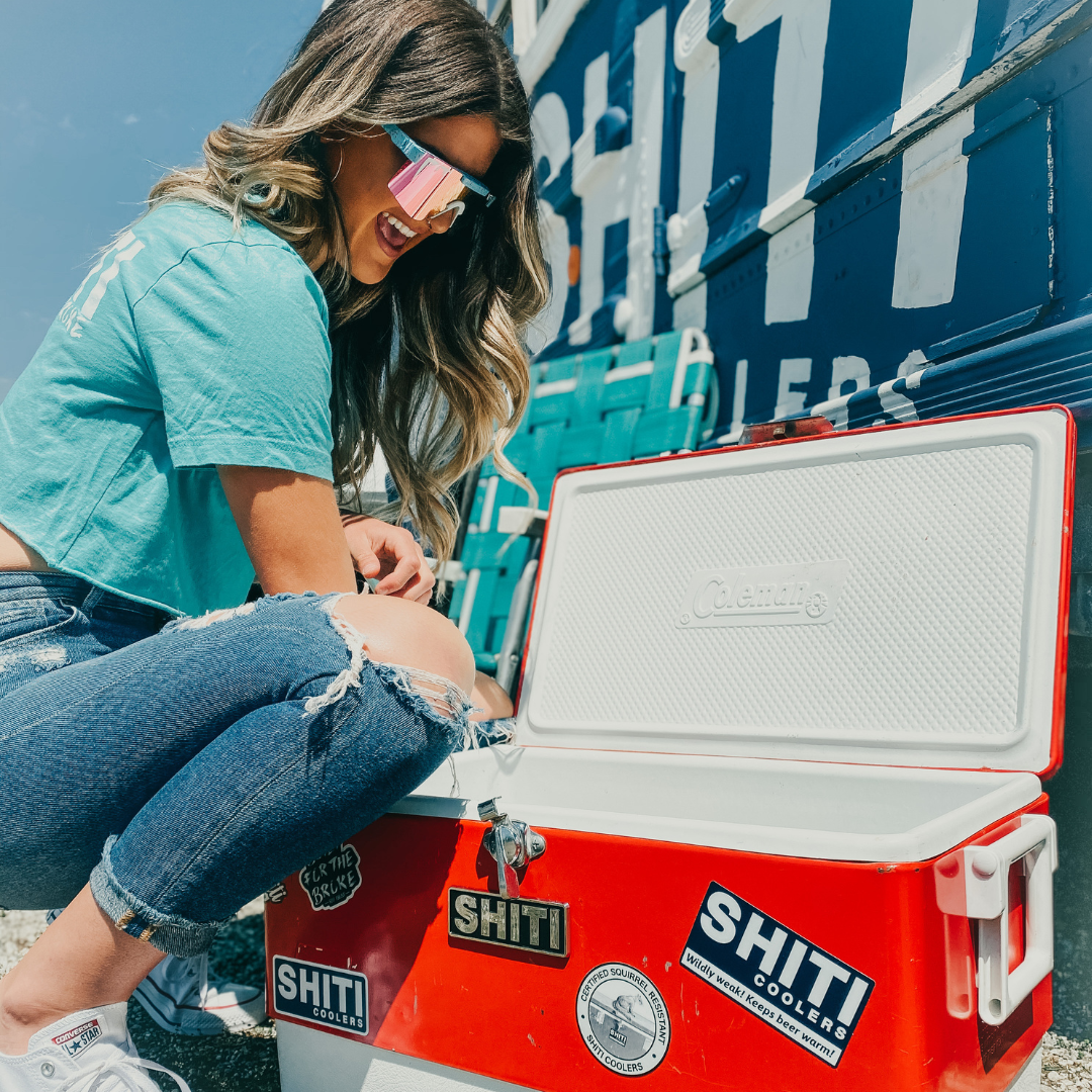 A woman in sunglasses opens a red cooler with stickers, showcasing the Chrome Brand Plate and enjoying a sunny day.
