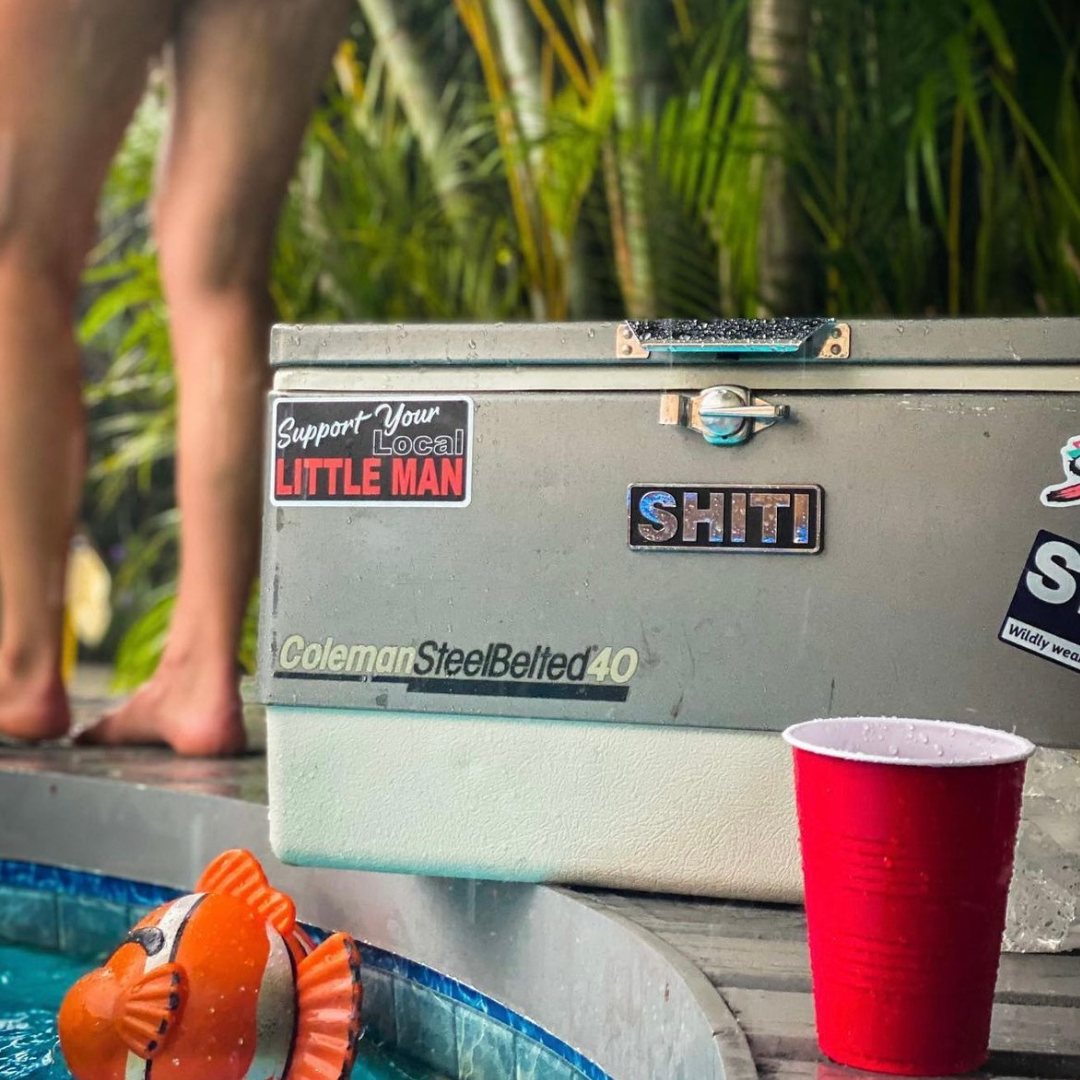 Chrome brand plate on a cooler with stickers, beside a pool and a red cup, creating a fun party atmosphere.