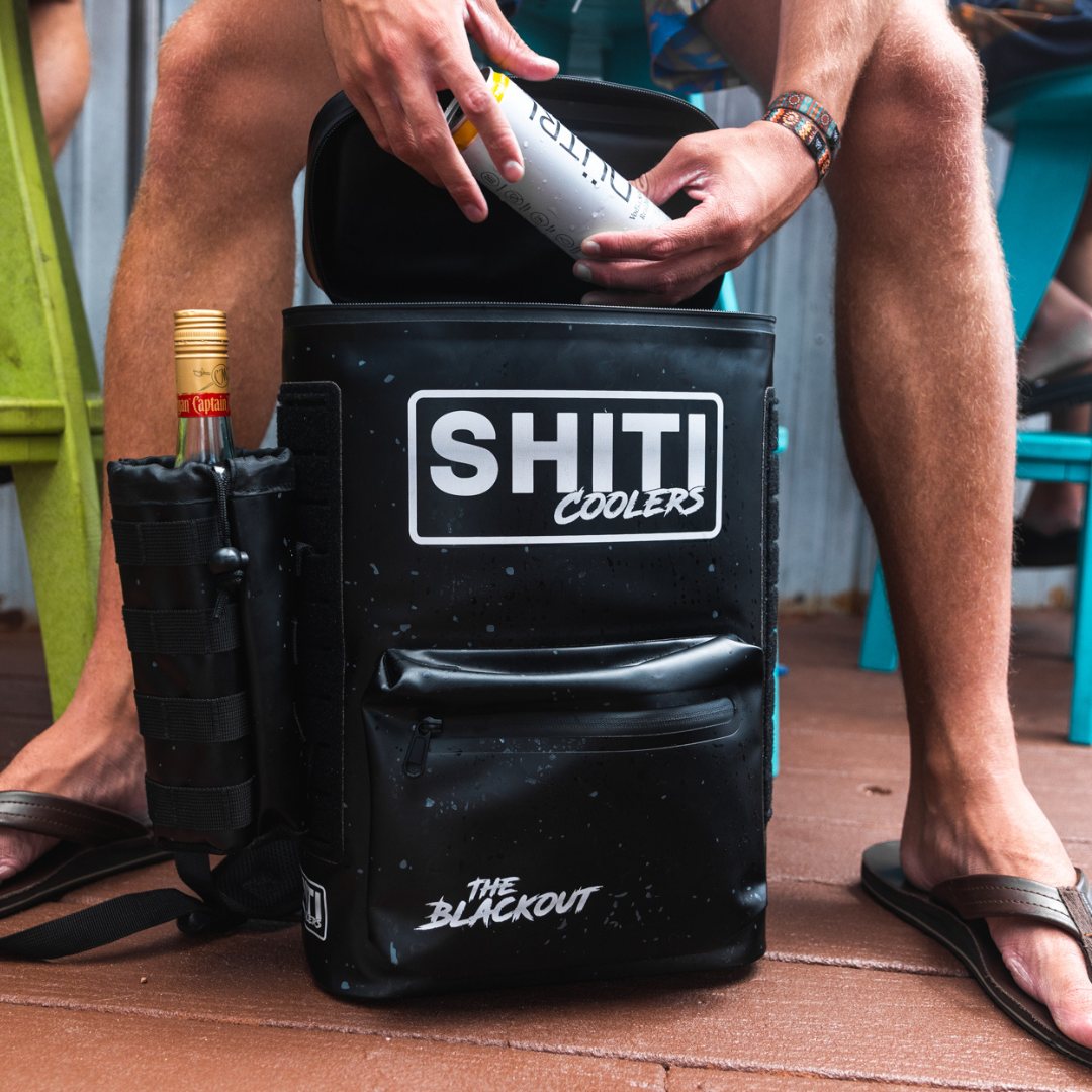 A person sitting on a deck is using the Blackout Drink Sleeve Attachment on a cooler, reaching for a beverage inside.