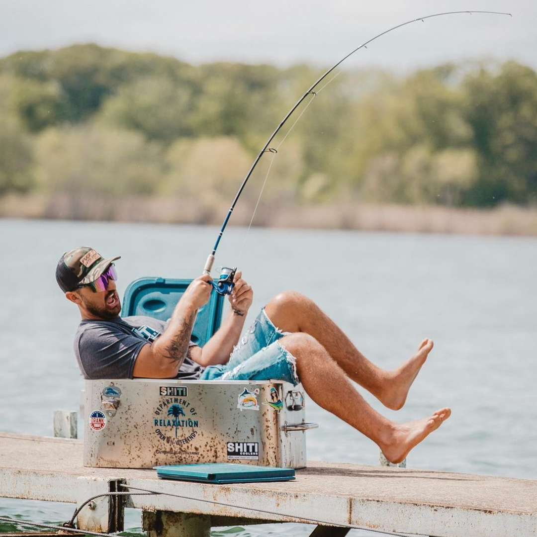 A man relaxes on a fishing cooler by the water, casting his line while enjoying a sunny day outdoors.