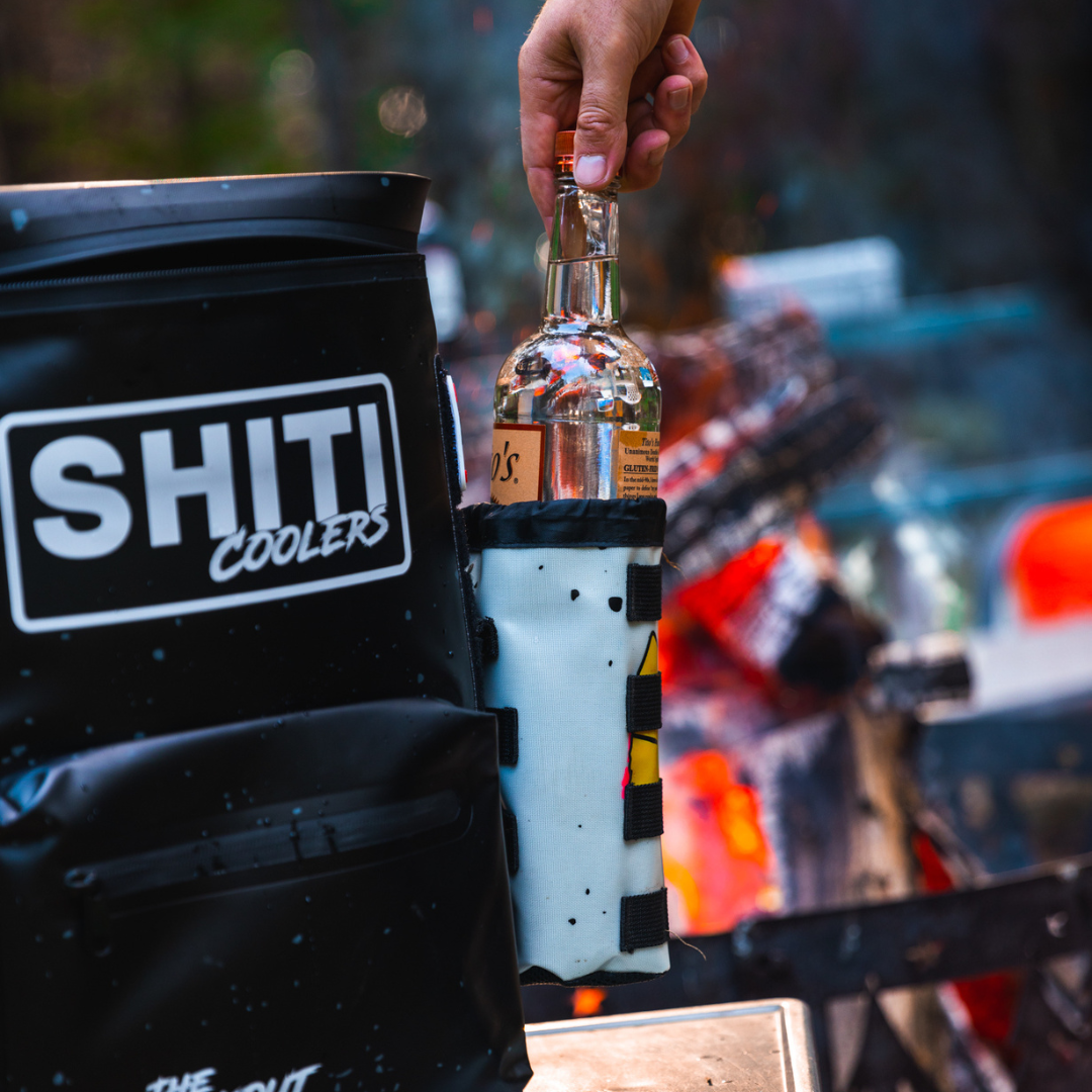 A person holds a bottle while attaching The Boozer Drink Sleeve to a SHITI Coolers bag near a campfire.